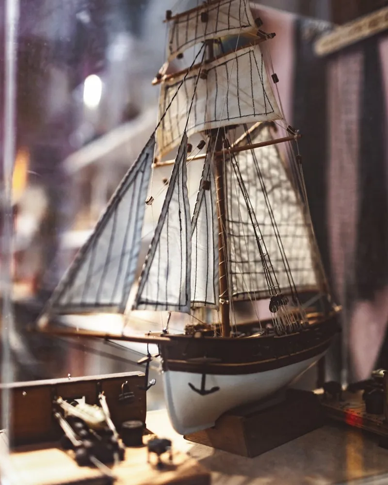 Maqueta de goleta con velas, mástiles y cabos finos expuesta dentro de una vitrina de cristal sobre su peana